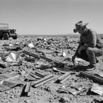 A 1947 vintage style black and white photo of a cowboy kneeling in a vast debris field of metallic UFO wreckage in the New Mexico desert.