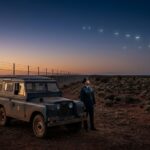 A 1960s RAAF military officer stands in the Australian outback looking up at glowing UFO lights near a prohibited area.