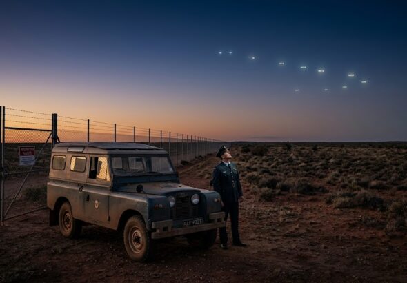 A 1960s RAAF military officer stands in the Australian outback looking up at glowing UFO lights near a prohibited area.