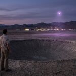 A radiation safety officer looking over a nuclear crater toward the lights of Area 51.